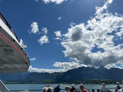 The image captures a picturesque scene from a boat on Lake Thun, Switzerland. In the foreground, the white and red structure of the boat's deck is visible, with a railing and several passengers seated on benches. The passengers, a mix of individuals, are looking out towards the lake and mountains.

The lake stretches across the middle ground, its calm, blue waters reflecting the bright sky. In the distance, a range of lush green mountains rises dramatically, their peaks dotted with snow in some areas. A small town or village can be seen nestled along the lakeshore, with buildings and trees adding to the scenic vista. The sky is a vibrant blue, filled with scattered white clouds that cast shadows on the landscape below, indicating a sunny day with some cloud cover. The overall impression is one of a serene and beautiful day in the Swiss Alps.