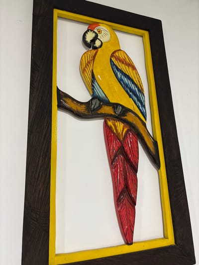 The image displays a framed, three-dimensional relief carving of a vibrant parrot, positioned against a white wall. The carving depicts a parrot with a predominantly yellow body, blue and yellow wing plumage, and distinct red tail feathers. Its head is yellow with a prominent black beak and a white area surrounding a black eye. The parrot is depicted perched on a dark brown branch, which is also part of the relief. The carving is set against a flat white background within the frame. The frame itself features a narrow yellow inner border and a wider, dark brown outer border, exhibiting visible wood grain. No individuals, actions, or interactions are present in the scene. The specific location within the city of Quimbaya, Colombia, is not ascertainable from the visual evidence provided.