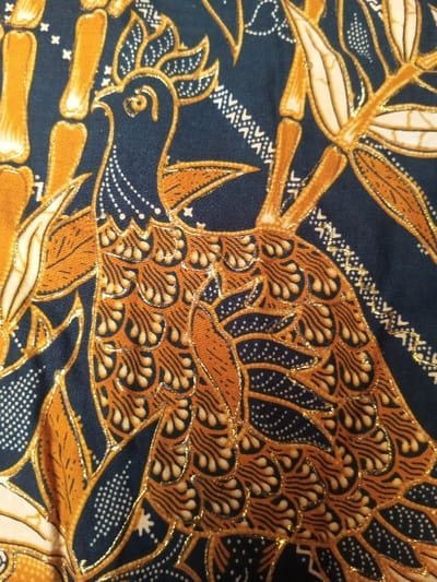 The image showcases a detailed batik pattern. A stylized bird figure with ornate feather details dominates the scene, colored in hues of brown and gold. The background is a dark blue, adorned with botanical elements resembling bamboo and leaves. Fine gold lines outline the shapes, adding a touch of elegance to the traditional design. Various small geometric patterns embellish the background, completing the artistic composition.