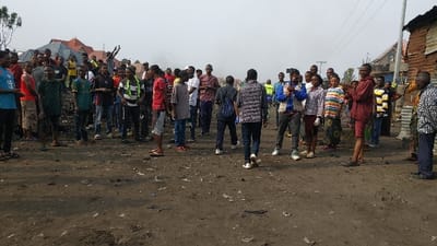 This image depicts approximately 40 to 50 individuals gathered on an unpaved dirt ground within a residential area of Gisenyi, Rwanda. The ground is littered with scattered debris. The individuals, primarily young adults, are casually dressed and standing in a loose formation, generally facing towards the center or left of the frame. One person in the foreground on the right, wearing a blue jacket, holds a smartphone horizontally, appearing to record. In the background, residential structures range from corrugated iron shacks on the right to more substantial buildings with tiled roofs, including one prominent orange-brown roof. Overhead power lines are visible on the upper right. A faint plume, possibly smoke or dust, is present in the mid-distance on the right. The sky is uniformly light gray, indicative of overcast or hazy conditions. The gathering appears observational, with no significant interactions among the larger group overtly discernible.
