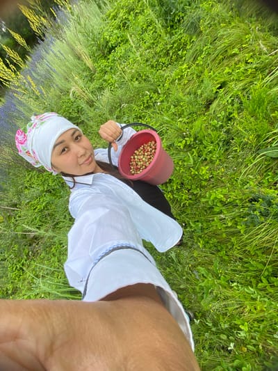 A young woman is depicted in a selfie, holding a red bucket filled with wild strawberries. She is wearing a white long-sleeved shirt, a white headscarf with a floral pattern, and dark pants. The setting is outdoors, in a field of tall green grass and wildflowers, with some purple flowers visible in the background. The lighting suggests it is daytime, likely a sunny day, as there are no shadows visible. The woman appears content, with a slight smile on her face, and is looking towards the camera. The image was taken in Maloarslangulovo, Russia.