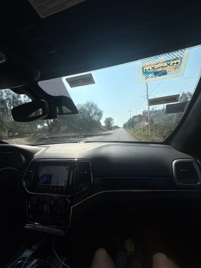 A view from inside a vehicle, possibly a car, looking out onto a road in Santiago Tlautla, Mexico. The dashboard and windshield frame the view. A navigation system is visible on the center console. Two stickers are affixed to the top of the windshield on the driver's side. The road leads into a landscape with trees and utility poles. Part of a person's legs and a green object are visible on the right side of the frame. The sky is bright with sunshine.