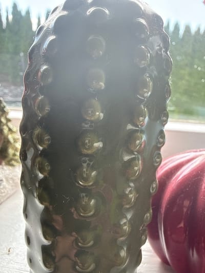 This is a close-up, indoor photograph of a dark, bumpy, candle-like object, possibly made of wax or resin, positioned in front of a window. The object is cylindrical and features a textured surface with raised, rounded protrusions. To the right of the main object, a portion of a shiny, ribbed, red object, likely another decorative item such as a pumpkin, is visible. In the background, through the window, blurred greenery and what appears to be a bright sky can be seen. The lighting suggests it is daytime. There are no people or animals present in the image. The location is stated to be in Ridabu, Norway.