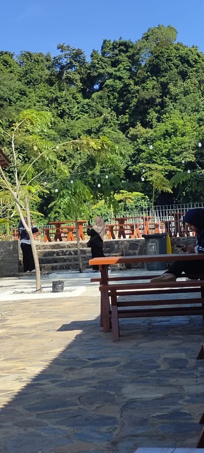This is an outdoor scene in Lamseunia, Indonesia, featuring a seating area amidst lush greenery. In the foreground, a person is seated on a wooden bench, looking down at a mobile phone. The bench is made of dark wood and is positioned on a stone-paved area. To the left and in the mid-ground, two individuals are standing, one in a black outfit with a headscarf, and another in a white top and black pants, also with a headscarf. They are near a tree adorned with white string lights. Behind them, several wooden picnic tables are arranged on a stone patio, separated by a white fence. The background is dominated by a dense forest with a clear blue sky above. The overall impression is a tranquil, natural setting, possibly a park or a recreational area.