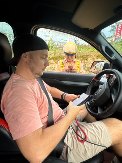 Here is a detailed description of the image:
An image taken from inside a car shows the car's driver being pulled over by a traffic officer in Quản Bạ Commune, Vietnam. The driver is a Caucasian man wearing a pink shirt, a black baseball cap, and a seatbelt. He is holding his cell phone and appears to be looking at it. Through the car window, the traffic officer can be seen. He is wearing a yellow helmet and a traffic officer's uniform. He is also holding a cell phone. The environment outside of the car shows local signage in Vietnamese. The presence of the uniformed official interacting with the person in the car suggests the potential enforcement of traffic regulations.
