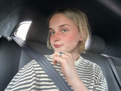 A young person with fair skin and light blonde hair is in the back seat of a vehicle, secured with a grey seatbelt. They are wearing a striped shirt and a ring on their finger. A portion of the vehicle's rear window and interior are visible. The location in Kelowna, Canada is not discernible from the image.