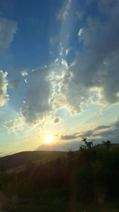 This image captures a sunset over a hilly, vegetated landscape in Zlatna Panega, Bulgaria. The sun, a bright orb of light, is positioned near the horizon, casting a warm glow that illuminates the sky and clouds. The sky is a vibrant mix of blues and oranges, with dramatic cloud formations. Rays of light are visible emanating from behind the clouds, creating a serene and ethereal atmosphere. The foreground is dominated by dark silhouettes of trees and bushes, indicating a natural, perhaps rural, setting. The overall impression is one of natural beauty and the tranquil end of the day.
