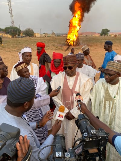 Several men, primarily of African descent, stand outdoors in traditional attire. Microphones from media outlets are held up to a man in a light-colored outfit. In the background, a bonfire burns intensely. The setting appears to be a field with buildings in the distance under a hazy sky. The men seem to be the focus of a press event or interview with the fire adding a dramatic backdrop to the scene.