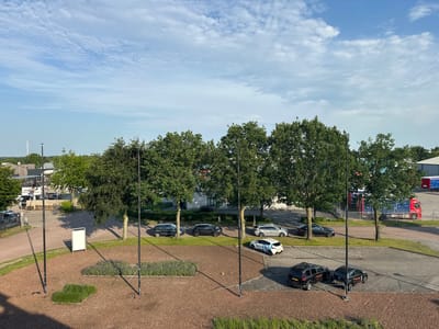 The image depicts an outdoor scene with a bright, partly cloudy sky. In the foreground, there is a section of dark brown ground cover, possibly wood chips, with patches of green grass and low-lying green plants. Two tall, black lampposts are visible. Further back, a paved area with parking spaces is present.

Several cars are parked:
* Two dark SUVs are parked side-by-side, both displaying the logo "Payned Partn...". One appears to be a Volvo XC90.
* A black sedan is parked to the left of the SUVs.
* Further up the road, a white car with blue racing stripes is parked.
* Two other cars are visible further back, one a silver SUV and another a dark sedan.

Trees with green foliage line the area, casting shadows. Beyond the trees, buildings and industrial structures are visible, including what appears to be a warehouse or factory. A large red truck with a blue trailer is parked in the distance. A white sign on a stand is positioned on the brown ground cover.

The scene is located in Staphorst, Netherlands, as indicated by the context. The overall impression is of a commercial or industrial area on a clear day. No people are visible in the image.