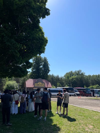 A group of people, appearing to be tourists, are gathered in an outdoor setting. In the background, a small wooden building with a red roof displays a sign in Cyrillic script that reads "СУВЕНІРЫ" (Souvenirs). Several buses are parked in a lot behind the building, suggesting a tourist destination or rest stop. The area is surrounded by trees and greenery, with a clear blue sky overhead. The people are casually dressed, and some are looking at their phones or interacting with each other. One individual is holding a small object, possibly a souvenir. The ground is covered with grass and gravel.