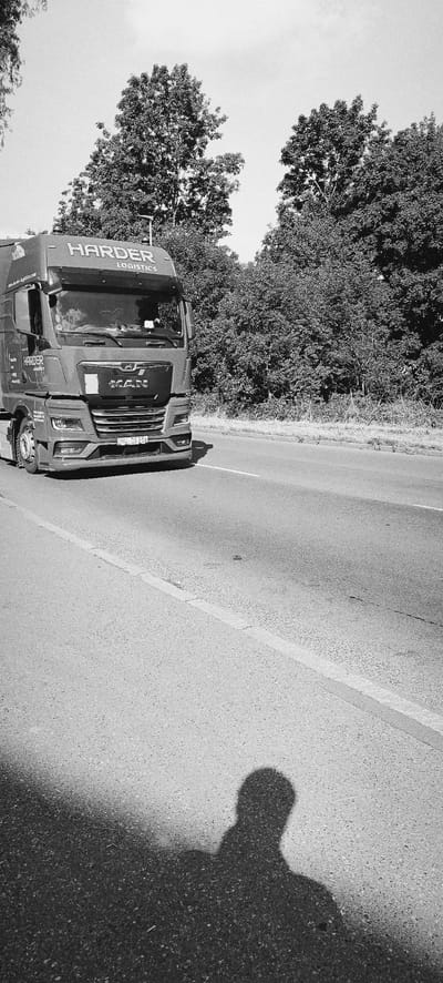 A black and white, eye-level, medium shot shows a MAN semi-truck with "HARDER LOGISTICS" written on its side, driving on a road. The truck is a dark gray color. The truck's front grill is clearly visible, with the "MAN" logo. The license plate reads "NU DS 556." The truck is traveling on an asphalt road with a white line dividing the lanes. Trees and foliage line the right side of the road, and the sky is visible in the background. A shadow of a person holding a camera is cast on the road in the foreground, obscuring the bottom left corner of the image. The scene appears to be captured on a sunny day. The location is identified as Waiblingen, Germany.