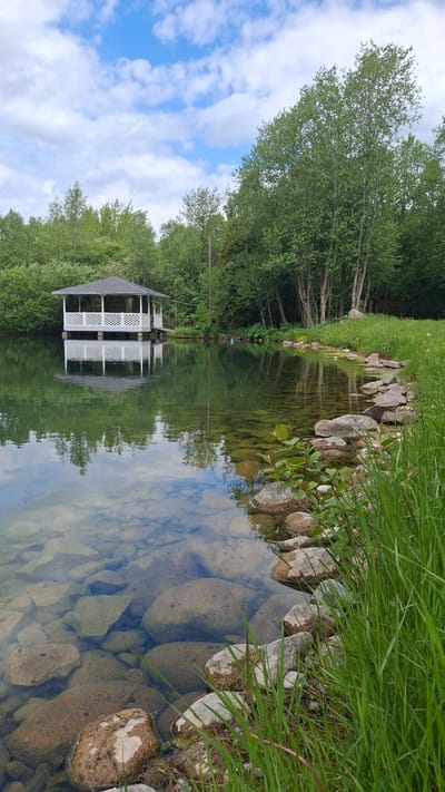 The image captures a serene outdoor landscape in Kipen, Russia, featuring a tranquil body of water, likely a pond or calm river, reflecting its surroundings. The water, clear enough to reveal submerged rocks and pebbles at its bottom, occupies a significant portion of the frame. Its surface provides a near-perfect mirror image of the bright blue sky dotted with white clouds and the dense, lush green trees lining the banks.
Along the immediate foreground and right side of the water, the bank is lined with various sized rocks and boulders, interspersed with vibrant green grass. On the opposite side of the water, a distinctive white gazebo with a dark, possibly gray, roof stands amidst a verdant backdrop of numerous trees. The gazebo's reflection is sharply defined in the still water.
The scene depicts a pleasant, bright daytime, likely late morning or early afternoon, under partly cloudy skies. No people or specific activities are visible, emphasizing the peaceful and undisturbed nature of the environment. The overall impression is one of natural beauty and tranquility in a well-maintained setting. There is no visible text in the image.
