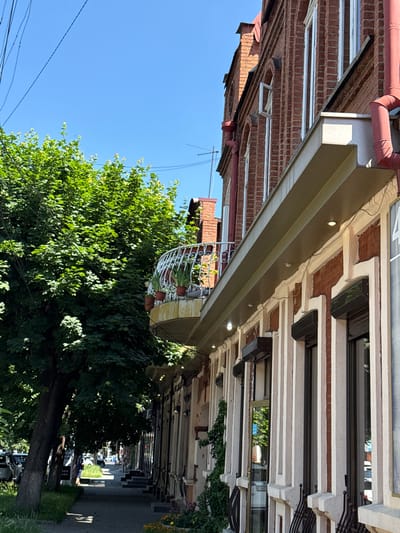 The image depicts a street scene in the city of Verkhnyaya Salda, Russia, on a bright, sunny day. In the foreground, a red brick building with a white ornate balcony adorned with potted plants stands prominently. The building features large windows, some with dark shutters, and decorative cream-colored vertical trim. A red pipe runs vertically along the side of the building. To the left of the building, a lush green tree with dense foliage casts shadows on the sidewalk. A paved sidewalk runs along the street, with cars parked on the left side and a line of shops or businesses visible further down the street. The sky is clear blue. There are no people clearly visible in the scene.