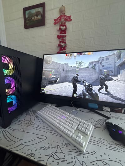 This image depicts a gaming setup in a room within a residence in Polkovnik Diakovo, Bulgaria. The central focus is a computer monitor displaying a first-person perspective from the video game *Counter-Strike: Global Offensive*. The game scene shows an outdoor urban environment with stone structures, an archway, and buildings; two in-game characters, dressed in tactical gear, are visible, one holding a rifle, and the player character holding a distinctive black and white patterned pistol. A military vehicle is partially visible through the archway. Game interface elements, including player statistics, a timer (0:04), scores (0-0), in-game money ($800), and a minimap, are present on the screen. The monitor also displays a faint "Activate Windows" watermark.
To the left of the monitor is a black desktop computer tower, featuring a meshed front panel with three vertically stacked RGB cooling fans displaying a rainbow color gradient. In front of the monitor and tower, a white gaming keyboard with visible "GENESIS" branding and white keycaps with some RGB backlighting rests on a large white mousepad. The mousepad features black line art illustrations of marine life, including a whale and fish, alongside stylized mountain landscapes. To the right of the keyboard, a black Logitech G gaming mouse with a blue illuminated logo is positioned on the same mousepad.
The background wall is covered with white, brick-patterned wallpaper. A small, framed painting depicting a rural house with a thatched roof and trees hangs on the upper left portion of the wall. To the right of the painting, a red and white "MERRY CHRISTMAS" banner hangs above a vertical string of four red and white Santa Claus figures.