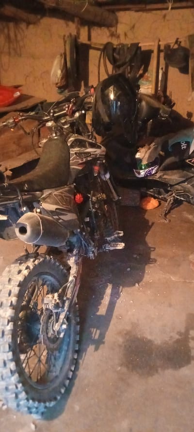 The image depicts a dirt bike parked indoors, likely in a garage or workshop, given the concrete floor and the wall in the background. The bike is the primary subject, with its front wheel visible on the left side of the frame. It has knobby tires, suggesting it's designed for off-road use. A black helmet rests on the handlebars, and another helmet with purple and white detailing is placed on the seat. The scene appears to be dimly lit, possibly indicating it's nighttime or the area has limited lighting. There are some items hanging on the wall in the background, including what looks like bags and possibly tools. An orange object is visible on the floor near the rear of the bike. The overall impression is one of a utilitarian space where a dirt bike is stored or maintained. There is no visible text in the image. The location context provided is Бетқайнар, Kazakhstan.