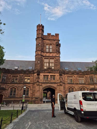 The image depicts the exterior of a large, gothic-style building constructed of reddish-brown stone. The building features a prominent tower with decorative stonework, arched windows, and a steep-pitched roof with skylights. In front of the building, a white van is parked with its driver's side door open. A person wearing a dark uniform with orange stripes on the pants stands beside the van, facing the building. Another person is partially visible inside the van. The foreground shows a paved walkway and a grassy area enclosed by a low fence. A bicycle is parked to the left of the pathway. The scene appears to be set in a university campus environment, consistent with locations in Princeton, New Jersey, USA, known for its collegiate gothic architecture. The sky is a clear blue with a few wispy clouds.