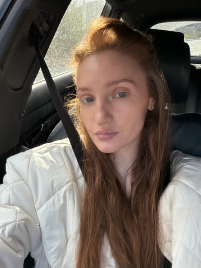 A woman with light skin and reddish-brown hair is shown in a close-up, slightly tilted view. She is wearing a white quilted jacket and a seatbelt is visible across her chest. The background indicates she is in a car. The location in Donji Ulići, Montenegro, is not discernible from the image.