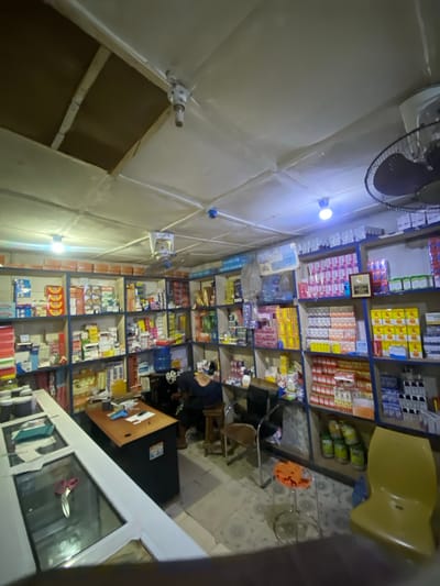The image depicts the interior of a pharmacy in Jere South, Nigeria. The pharmacy is well-stocked with numerous shelves filled with various medicines and health products. A person, likely a customer or employee, is seated at a desk in the middle of the room, looking down. The lighting in the pharmacy appears to be a mix of overhead fluorescent lights and possibly some natural light from an unseen source, creating a somewhat dim and uneven illumination. The overall impression is of a functional, albeit somewhat cluttered, retail space.