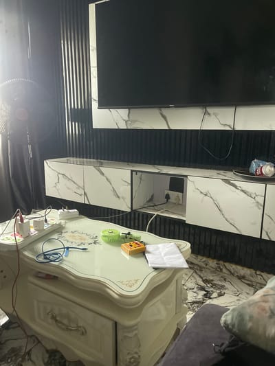 A fan is positioned to the left of the frame, partially obscuring a black curtain and a vertical slat wall. A large black television is mounted on the wall above a white and grey marble-patterned media console. The console has multiple compartments, one of which is open, revealing electrical outlets and plugs. In the foreground, a light-colored, ornate coffee table with a glass top holds several items, including a white power strip with multiple plugs and devices, a small green fan, a box of matches, and a folded piece of paper. The floor appears to be covered in a marble-patterned tile. The scene is indoors, consistent with a living room. The image does not contain explicit information about the city of Naze, Nigeria, or any specific individuals.