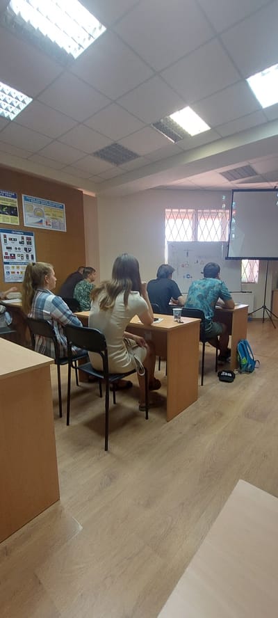 The image depicts an indoor scene, likely a classroom or lecture hall, with several individuals seated at desks. The room features a suspended grid ceiling with fluorescent lighting panels. The walls are light-colored, and one wall displays several posters with text and graphics. A projector screen is set up at the front of the room, displaying content.

In the foreground, on the left, a young woman with blonde hair tied back is seated at a desk, wearing a plaid shirt. To her right, another woman with long brown hair is seated, wearing a light-colored dress. Further to the right, a person with dark hair is seated at a desk, wearing a patterned shirt, followed by another individual in a dark shirt. On the far right, a person wearing a blue and green patterned shirt is seated.

The desks are made of light wood, and chairs are simple black metal frames with dark seats. On the desks, there are various items including cups, a phone, and papers. A blue backpack and a black cap are visible on the floor near the desks on the right side of the image. The floor is covered with light-colored laminate flooring. The overall impression is that of a learning environment, possibly during a presentation or lesson. The specific location in the city of Tetiiv, Ukraine is not determinable from the visual information provided in the image.