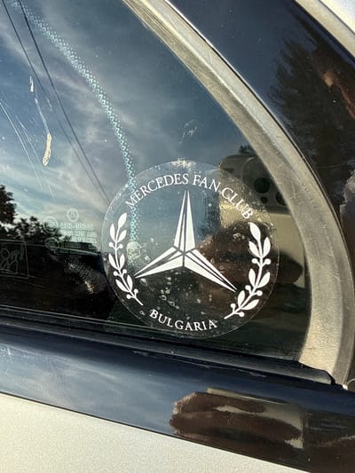 This is a close-up, eye-level, outdoor photograph of a sticker on a car window. The sticker is circular and black with white text and graphics. The text reads "MERCEDES FAN CLUB" in an arc at the top and "BULGARIA" at the bottom. In the center of the sticker is a white three-pointed star, the Mercedes-Benz logo, enclosed by what appears to be laurel branches. The sticker is slightly dirty and appears to have some water spots on it. To the left of the sticker, on the window, are some small, faint markings, and what appears to be a certification mark. The car appears to be parked outdoors, with trees and sky visible in the reflection on the window. The bottom of the image shows the dark blue paint of the car door. This sticker suggests the car is associated with a Mercedes-Benz fan club and is located in Bulgaria.