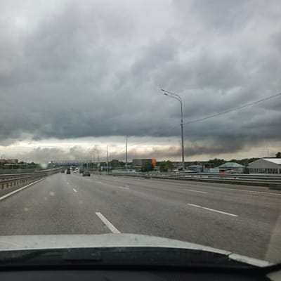 This media file depicts a view from a car traveling on a highway. The scene is characterized by a vast, overcast sky filled with dark, heavy clouds, suggesting an impending storm or a generally gloomy day. The highway stretches ahead, with multiple lanes and guardrails visible. Several cars are on the road, indicating active traffic.

In the distance, on the left side of the highway, there are buildings and a dense arrangement of trees. On the right side of the highway, more industrial-looking buildings and structures are visible, along with trees. A prominent feature in the middle ground, across the highway, is a large plume of dark smoke rising into the sky, obscuring the horizon. This smoke appears to be originating from the industrial area.

Streetlights are spaced along the sides of the highway, some of which are visible as curved metal poles with lights. There are also road signs, including a blue sign with white text and an arrow pointing to the right, which reads "МИХАЙЛОВ" (Mikhailov).

The overall impression is one of a potentially hazardous or concerning situation due to the dark sky and the large smoke plume, juxtaposed with the ordinary activity of highway traffic. The time of day is not explicitly clear but the overcast conditions suggest it could be daytime. The location is identified as Oktyabrskiy, Russia. There are no visible people in the scene, and the focus is on the landscape and the ongoing journey.