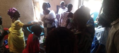 The image captures an indoor gathering of people, predominantly women and children, likely in Ohafia, Nigeria. The room appears somewhat crowded, with bright natural light entering from a doorway or window in the background, creating a backlit effect for some figures.
In the left foreground, a woman wearing a vibrant yellow dress and a distinctive purple head tie (gele) is shown in motion, with her right arm raised. She appears to be dancing, gesturing, or actively participating in an event. The dress features subtle embellishments, possibly embroidery, on the sleeves and hem. Next to her, a child in a red top is partially visible in the lower right corner.
Further back and towards the center, other individuals are present. One woman wears a white blouse with short puff sleeves and a patterned skirt, while others are seen in various light and dark colored attire. Light blue plastic chairs are visible in the background, suggesting seating arrangements, though many individuals are standing. Small bottles are scattered on the floor in the foreground. The walls of the room have a textured, off-white appearance.
The scene conveys a sense of a lively social event or community gathering during the daytime. The attire and head ties worn by the women are characteristic of Nigerian culture. No visible text is present in the image.