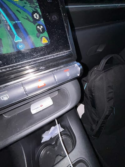 The interior of a vehicle is visible, including a navigation screen displaying a map with a route. A central console features a column of buttons with symbols for hazard lights, door locks, and window defrost. A small cubby adjacent to the buttons contains a white rectangular device with "VINCI Autoroutes" printed on it. A black bag is resting on the passenger seat. A charging cable is plugged in near the cup holders. Loose papers are also in the same cup holder area.