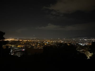 An elevated, wide-angle night scene captures the urban landscape of Gerakas Municipal Unit, Greece. The foreground consists of dark, silhouetted foliage and tree branches partially obscuring dimly lit building structures. The mid-ground and background reveal a dense expanse of city lights, predominantly yellow and white, indicating illuminated buildings and street networks stretching into the distance. A faint, blurred white light, possibly an aircraft, is visible in the sky above the city horizon towards the left-center. The sky above is dark and heavily overcast with multiple layers of clouds, obscuring most celestial features, though a single, faint star or distant light source is barely discernible in the upper left quadrant. No individuals or specific actions are identifiable within the frame.