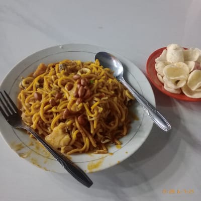A white plate containing a serving of yellow noodles with peanuts, seafood, and a brown sauce sits on a white table. There is a fork and a silver spoon next to the noodles. There is another plate with a red rim on the table holding several fried white crackers. The location is Kecamatan Panongan, Indonesia, at coordinates (latitude: -6.264, longitude: 106.525).