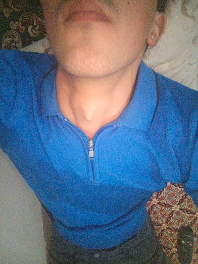 The image displays a close-up, angled shot of a person's upper body, focusing on the chest, neck, and lower part of the face. The person appears to be lying down or reclining, with their head tilted slightly backward.

The individual is wearing a blue, long-sleeved polo-style shirt made of a knitted or textured fabric. The shirt has a collar and a half-zip closure in the front, with a metallic zipper pull visible at the bottom of the open placket. A prominent Adam's apple is visible on the person's neck.

Above the shirt, the lower half of the person's face is visible, including the chin, jawline, and mouth area. There is light stubble or short facial hair on the chin and around the mouth. The lips are slightly parted. The right ear is partially visible on the right side of the frame, near the collar of the blue shirt.

At the very bottom of the image, a dark, solid-colored garment, possibly trousers or shorts, is visible. To the right of the person's body, a section of patterned fabric is visible. This fabric features an intricate, repeating geometric and floral design in various shades of red, orange, green, and cream, suggesting a blanket, cushion, or upholstered surface.

The background on the left side of the frame appears dark and textured, while the background on the right is lighter and less distinct. The overall lighting is soft, and the image has a somewhat grainy quality. No text is visible in the image.