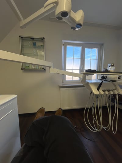The image depicts the interior of a dental examination room located in Monsheim, Germany, from a first-person perspective, looking forward from a seated position. The lower legs of a person wearing dark trousers and brown shoes are visible in the foreground. The room features white walls and a dark wooden floor. A large window, segmented into multiple panes by white frames, dominates the right side of the frame, admitting bright natural light and revealing a blue sky with faint clouds. A white radiator is positioned directly beneath the window.
To the left, a silver-framed glass display cabinet is mounted on the wall, containing various dental hygiene products, including bottles of Listerine mouthwash and packaging for GUM brand items. Above, a white, articulated dental examination light fixture with multiple lamp heads is suspended from the ceiling. A long, white, articulated arm extends into the mid-ground, likely part of the dental chair or unit's support structure.
To the right, a multi-component white dental unit is situated near the window. It features multiple white hoses with black tips hanging from its side, typical of dental instruments. On the unit's top surface, a dark folded cloth, a small silver object, and a white bottle labeled "NFS" are present. The "AIRFLOW" brand logo is visible on the main body of the dental unit. No other individuals or active procedures are depicted.