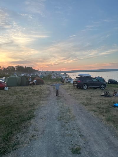 A wide-angle, eye-level shot captures a bustling campsite at dusk, likely near the city of Zhemchuzhny, Russia. The sky above is a canvas of soft blues and oranges, streaked with wispy clouds, suggesting the sun is either rising or setting over the adjacent body of water. A sandy, unpaved path runs down the center of the frame, leading the viewer towards a large gathering of tents and vehicles.
In the foreground, a young person stands with their arms outstretched, facing the viewer. They are wearing a light gray hooded sweatshirt and striped pants, and appear to be a child or young teenager. The path they stand on is well-trodden, indicating frequent use by campers.
Scattered across the grassy terrain are numerous tents of various sizes and colors, with some exhibiting signs of recent activity. Several vehicles are parked among the tents, including a dark SUV with a white roof box on top, a red car, and a black car. A small campfire pit, containing burnt wood, is visible on the right side of the path.
In the background, a dense line of trees forms a dark silhouette against the horizon. Beyond the trees, more tents and structures are visible, suggesting the campsite extends for a considerable distance. The calm water of a large lake or reservoir borders the campsite on the right side of the frame. The overall atmosphere is one of a lively outdoor gathering, possibly a festival or a large camping event.
