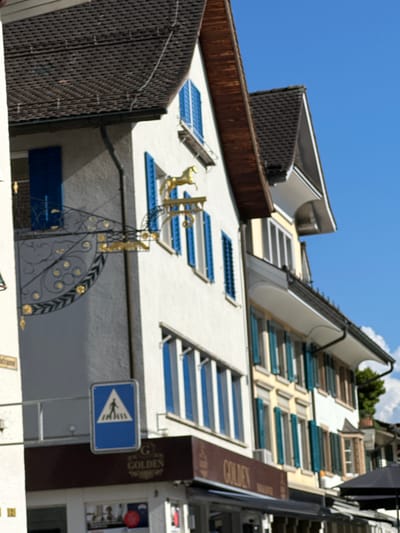This is a side view of a building in the city of Wald (ZH), Switzerland, under a clear blue sky. The building has multiple stories with white and yellow facades. The roofs are dark and tiled. Numerous windows are visible, many with closed blue shutters. A golden decorative element resembling a horse is mounted on the side of the building, above an ornate black metal sign bracket with gold accents. Below this, a blue pedestrian crossing sign is affixed to the wall. The ground floor of the building features signage with the word "GOLDEN" prominently displayed. An outdoor cafe setup with a black umbrella is partially visible in the lower right corner.