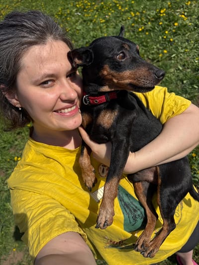 A woman is smiling up at the camera while holding a black and brown dog in her arms. The dog is wearing a red collar. The woman is wearing a yellow shirt and is laying in a grassy area with yellow flowers. The location is unknown and there is no indication of being in the city of Усть-Тунтор, Russia.