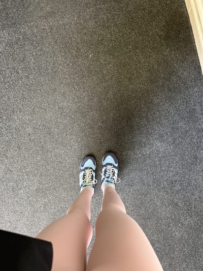 The image shows a person's legs and feet, wearing blue and white athletic shoes, standing on a gray carpeted floor. The perspective is from above, looking down at the person's feet. The scene is indoors, with a white trim visible on the right side of the image, possibly indicating a doorway or wall. The lighting suggests a well-lit indoor environment. No specific landmarks or details are visible to identify a location within the city of Carlow, Ireland.