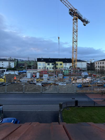 The image depicts a construction site located in Givisiez, Switzerland, captured during daylight hours under an overcast sky.
In the immediate foreground, the top surface of a reddish-brown corrugated roof structure is visible. Below this, an asphalt road with a double dashed white line extends horizontally. A dark blue vehicle is partially visible on the left side of the road, while a black metal fence and a patch of green grass are on the right.
The central focus is an active construction site, enclosed by temporary grey metal fencing. A prominent yellow tower crane is positioned on the right side of the site, its jib extending leftward over the construction area, with a heavy hook suspended from its cable. The site exhibits ongoing ground-level construction work, including concrete foundations, wooden formwork, exposed rebar, and multiple pre-cast concrete elements. Piles of excavated earth and gravel are distributed around the perimeter. A yellow excavator is partially visible on the left side of the construction zone. Two individuals, wearing orange high-visibility jackets and white hard hats, are present within the site, indicating construction personnel. One worker is positioned near the excavator on the left, and another is located towards the center-right amidst the concrete structures.
In the background, a row of multi-story residential buildings with dark roofs and varied light-colored facades (cream and yellow) stands directly behind the construction site. Further in the distance, additional residential structures, green rolling hills, and utility poles supporting overhead power lines are visible beneath the cloudy sky.