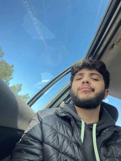 A male individual is depicted in a selfie taken from inside a vehicle in Al-Kiswa, Syria. The subject, a young adult male with dark, curly hair and a full, neatly trimmed beard, is positioned in the lower right portion of the frame, looking directly at the camera with slightly pursed lips. He is wearing a black quilted puffer jacket over a dark grey hooded sweatshirt with light green drawstrings. Above him, a clear blue sky with some faint linear marks and reflections is visible through an open sunroof. Green tree foliage is partially visible in the upper left quadrant, either reflected on the glass or visible through the sunroof. The vehicle's interior roof lining and the sunroof's structural frame are also visible.