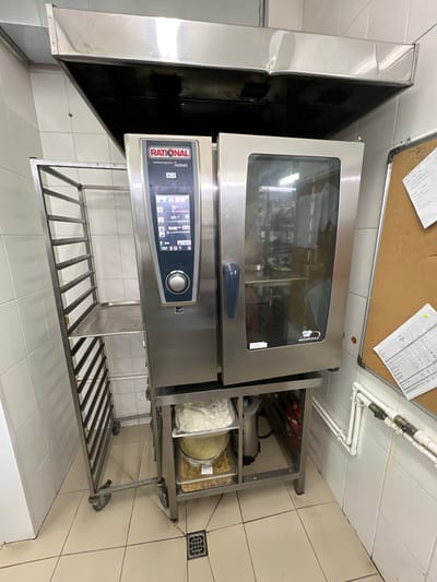 The image depicts a commercial kitchen scene. In the center, a large, stainless steel Rational combi-oven with a digital display is prominently featured. To its left is a multi-tiered stainless steel rack on wheels, designed to hold baking sheets or pans. Underneath the combi-oven, a metal shelf holds several food containers, one filled with white shredded material, and another with yellow granular material. The combi-oven and rack are positioned on a tiled floor. In the background, a stainless steel exhaust hood is mounted above the oven. The walls are covered in white tiles. To the right of the oven, a corkboard with papers is affixed to the tiled wall, and white pipes with valves are visible. The scene appears to be indoors, likely in a professional food preparation environment. The location provided, Plastunivska, Russia, suggests this is a commercial establishment within that region.