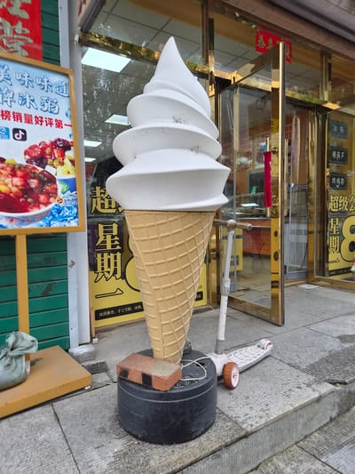 A large, white soft-serve ice cream cone sculpture is the focal point of this outdoor scene in front of a shop in Hunchun, China. The ice cream cone is on a black circular base, with a brick and a part of a scooter resting on or near it. A white kick scooter with pink accents is positioned to the right of the ice cream cone. To the left of the ice cream cone, a yellow sign with Chinese text and an image of a fruit salad is displayed. Behind the sign, a green corrugated metal shutter is visible. The shop entrance, with a gold-colored frame, is to the right of the ice cream cone, and through the glass doors, a display area with shelves and products can be seen. Several signs with Chinese text are posted on the shop's facade. The ground is paved with gray square tiles and concrete. A gray fabric bag is placed on a yellow platform to the left of the ice cream cone. The overall environment appears to be a commercial street.
