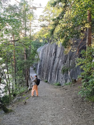 A person, viewed from behind, stands on a gravel path in Skriketorp, Sweden, holding a smartphone to take a photograph. They are wearing an off-white baseball cap, a light gray jacket, and bright orange pants. A black backpack is visible on their back. The path winds alongside a tall, sheer rock face on the right and dense green foliage with tall trees on the left. Sunlight filters through the leaves, casting dappled shadows. In the background, a body of water can be seen through the trees. The overall scene depicts a scenic outdoor environment, likely a hiking trail.