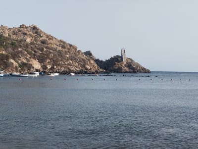 The image shows a coastal scene in the municipality of Águilas, Spain, featuring a rocky hillside with scattered vegetation and a prominent lighthouse-like structure on the right side. Several small boats are moored in the water along the coastline, and a series of red buoys are visible in the water. The sea is calm, with gentle ripples on the surface. The sky is clear and pale blue. The overall impression is one of a serene, sun-drenched coastal landscape.