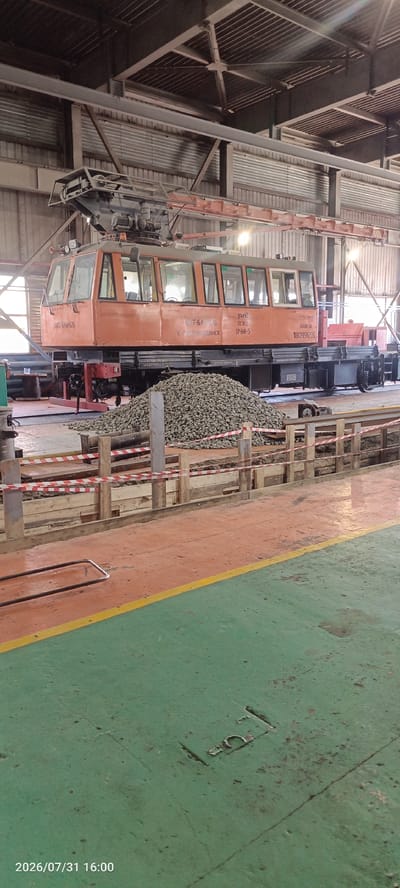 An orange and grey railway maintenance vehicle, possibly a crane or a ballast tamper, is parked inside a large industrial building. The vehicle is on tracks and has a pantograph extended to the ceiling. A large pile of grey ballast rocks is in front of the vehicle. The floor is painted with orange and green sections, separated by a yellow line. Red and white caution tape is visible around a section of the floor in the foreground, indicating a work area or hazard. The background shows the structure of the building, including metal beams and corrugated metal walls. The date stamp "2026/07/31 16:00" is present in the bottom left corner of the image. The location is identified as Severobaikalsk, Russia.