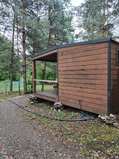 The image depicts a small, wooden cabin with a covered porch situated in a wooded area. The cabin is built on a foundation of stones and has a dark-colored roof. A black hose with blue connectors is visible running from the side of the cabin towards the foreground. The surrounding environment is characterized by tall pine trees and a gravel path, suggesting a rural or campsite location. The ground is covered with fallen leaves and some greenery. The overall scene is overcast, indicating a cloudy day. The location is identified as the city of Катунь, Russia.