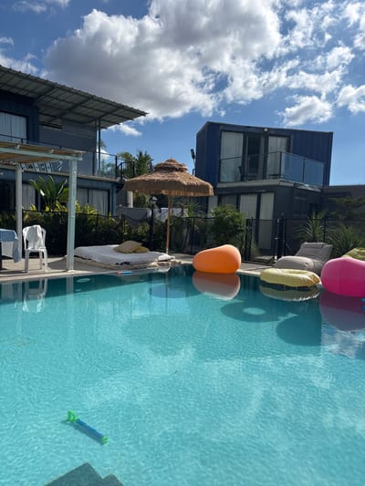 The image depicts an outdoor leisure area in Maale Yosef Regional Council, Israel, under a partly cloudy sky. In the foreground, a rectangular swimming pool with clear turquoise water occupies the lower half of the frame. Steps are visible beneath the water surface in the lower left corner, and a blue and green water toy floats partially submerged near the bottom center.
Around the pool, several objects are present. On the left side of the pool deck, a white plastic chair with a white garment draped over its back stands next to a table covered with a blue and white checkered tablecloth, beneath a white-framed canopy. Directly across the pool, a large, rectangular sunbed with white cushions and two light brown pillows is positioned on the ground. Adjacent to the sunbed, closer to the pool, a straw-thatched umbrella with a wooden pole stands upright.
To the right of the umbrella, a series of inflatable floats are either on the pool deck or partially submerged in the water: an orange, beanbag-shaped float in the pool, a grey beanbag-shaped lounge chair on the deck, a yellow inflatable ring float in the pool, and a bright pink beanbag-shaped float in the pool.
In the background, two multi-story buildings constructed from shipping containers are visible. The building on the left is dark gray/blue, featuring a flat metal roof overhang on its upper level and large windows or glass doors. The building on the right is dark blue on its upper level and dark gray/black on its lower level, also featuring large windows or glass doors and an upper-level balcony with a glass railing. Green foliage, including shrubs and palm-like plants, is present between the buildings and behind a black metal fence that separates the pool area from the structures. The sky is blue with scattered white cumulus clouds, and the scene is illuminated by direct sunlight, casting reflections on the water. No individuals are visible in the image.