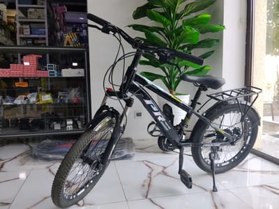 A black Prestige mountain bicycle is parked indoors, leaning on its kickstand. The bicycle has a black frame, black handlebars with "PRESTIGE" branding, and black grips. It features a black saddle, a rear rack, disc brakes, and knobby tires. The front tire is partially visible and appears dusty. Behind the bicycle, a large green plant in a white pot is situated next to a window. In the background, shelves stocked with various items, including boxes and what appear to be car parts, are visible. The floor is tiled with a marble pattern. The scene is brightly lit, suggesting an indoor commercial space, possibly a bicycle shop or a retail store selling automotive or mechanical parts in Yovon, Tajikistan. There are no people present in the image.
