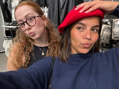 Two young adult females are depicted in a selfie taken inside a retail clothing store in Philadelphia, United States. The woman on the left has long, wavy reddish-blonde hair, fair skin, and wears black-rimmed eyeglasses, small gold hoop earrings, a layered gold chain necklace with a heart-shaped charm and an initial pendant, and a black sleeveless top. She is puckering her lips and looking directly at the camera. The woman on the right, positioned closer to the camera and likely holding it, has medium skin tone, dark brown shoulder-length hair, and wears a red beret, which she is holding with her right hand. She is dressed in a dark blue long-sleeved sweater and is also puckering her lips, looking slightly to the right of the camera. The background consists of typical retail store fixtures, including clothing racks displaying various garments such as white t-shirts, black tops, and a black faux leather jacket or vest. Merchandise such as handbags, some with a snakeskin pattern, are visible on shelves behind the woman on the right. A mannequin leg wearing black sheer tights is partially visible on the far left. The lighting is bright and even, characteristic of an indoor commercial space.