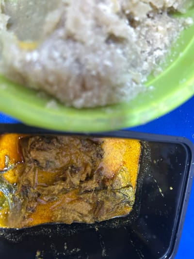The image displays a top-down view of a meal in Ilishan, Nigeria. In the upper portion, a light green bowl contains a light beige, granular, semi-solid food substance, possibly a staple like pounded yam or fufu. Directly below it, a black rectangular container holds a stew or soup. This stew features a prominent reddish-orange liquid base and contains dark brown, fibrous solids, consistent with cooked meat or fish, along with visible green leafy components. The containers rest on a vibrant blue surface. No individuals or specific actions are depicted.