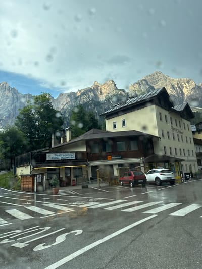 The image depicts a street scene in San Vito di Cadore, Italy, with a prominent view of the Dolomite mountains in the background. The foreground features a wet asphalt road with white crosswalk markings, indicating a pedestrian crossing. Two cars, a red compact car and a white SUV, are parked in a lot adjacent to a building. The building appears to be a multi-story establishment, possibly a hotel or apartment building, with a cream-colored facade and dark-pitched roofs. A sign above a ground-level storefront reads "Gastronomia Pane Minimarket." Another sign further down the building says "BAR ANTELAO." Green trees are visible between the buildings and the mountain backdrop. The sky is overcast with some patches of blue visible. The presence of water droplets on the camera lens suggests it may have been raining or the image was taken through a wet window.