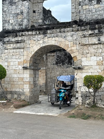 In Oslob, Philippines, a turquoise scooter with a blue patterned canopy is parked near a weathered stone archway. The scooter is a three-wheeled vehicle, with a sidecar attached, and appears to be carrying some sort of cylindrical containers. A person is seated on the scooter, looking towards the camera. The archway is part of an old stone structure, possibly a ruin, with visible signs of age and decay. The stones are rough-hewn and irregular, with patches of dark discoloration suggesting weathering or moss growth. A small, well-maintained green tree stands to the right of the archway. In the foreground, a paved road leads towards the structure. The sky above is overcast.